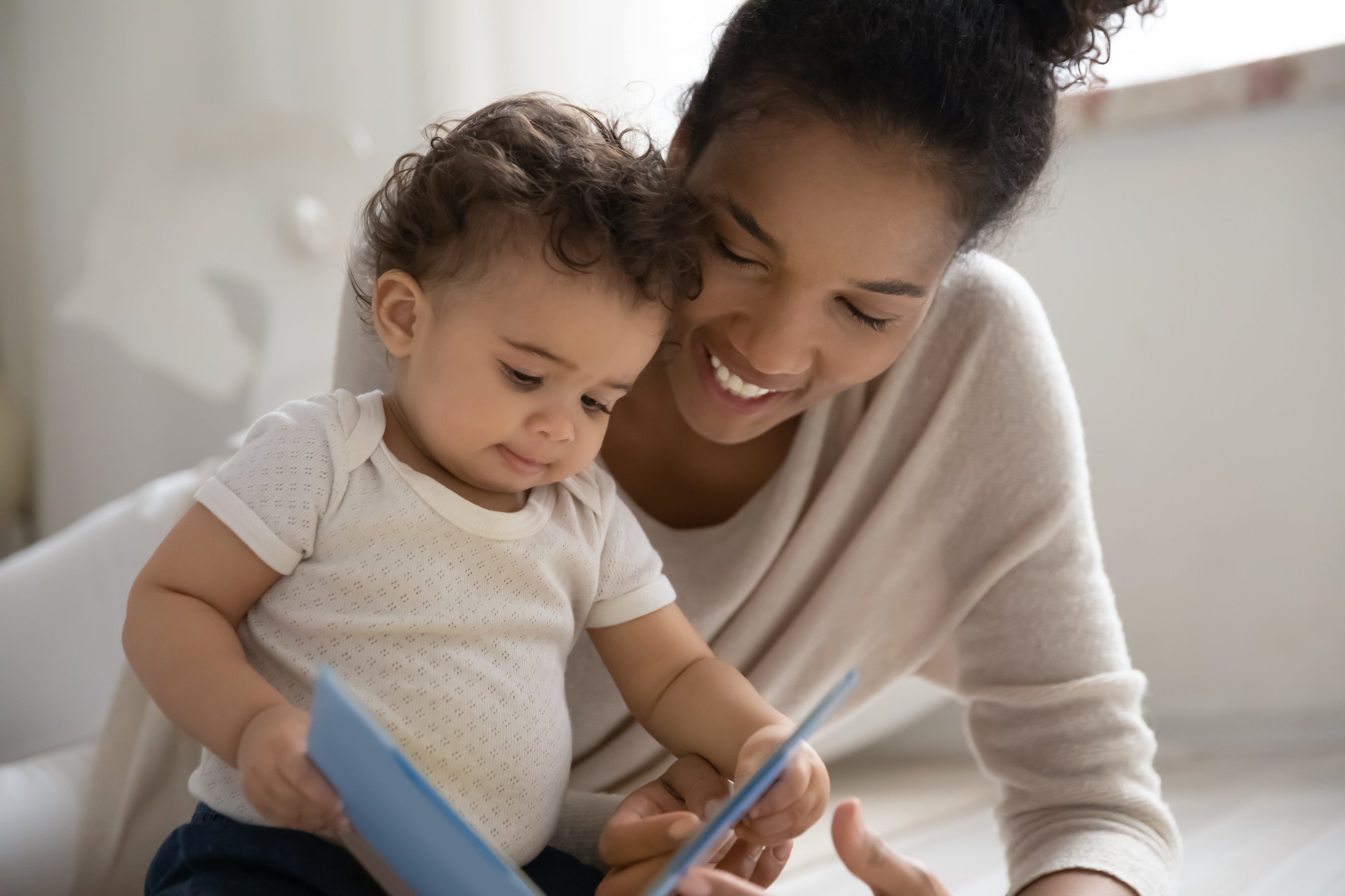 A mother and her infant sit on the floor, smiling while reading a book together - creating a special bonding moment with a kids book club designed for baby's first year.