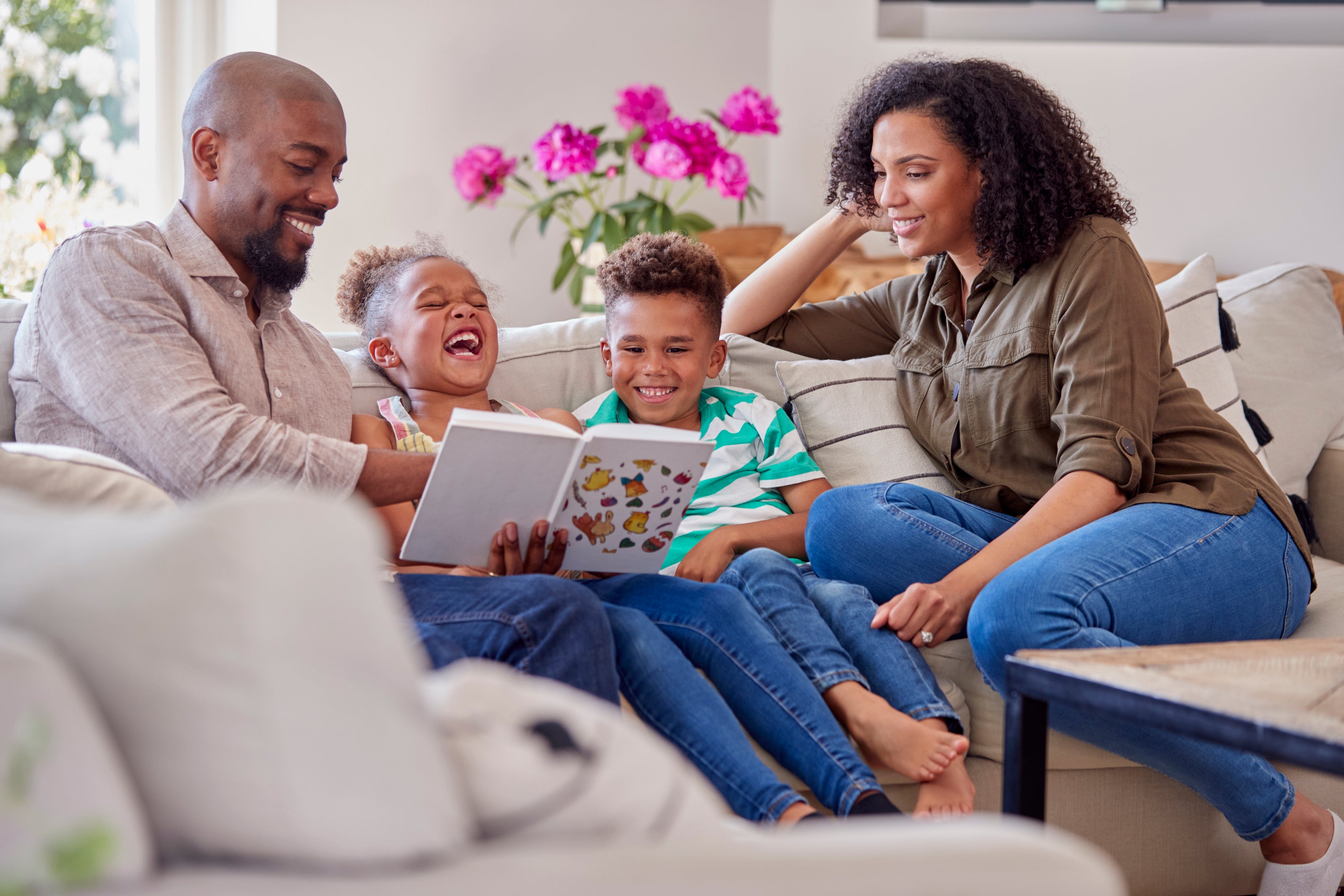 A happy family - mom, dad and two children - sits on a cozy sofa, laughing and reading a book together. Discover the best kids book club for adventure-filled stories that bring families closer!